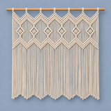 A cream macrame wall hanging with intricate woven geometric patterns and long fringe, suspended from a light natural wood rod against a solid dusty blue-gray background.