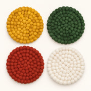 Four round felt pom-pom coasters arranged in a 2x2 grid on a white background, featuring yellow, green, red, and off-white colors.
