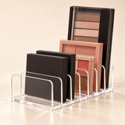 Clear acrylic makeup organizer stand holding several eyeshadow palettes in black and warm neutral shades against a soft beige studio background.
