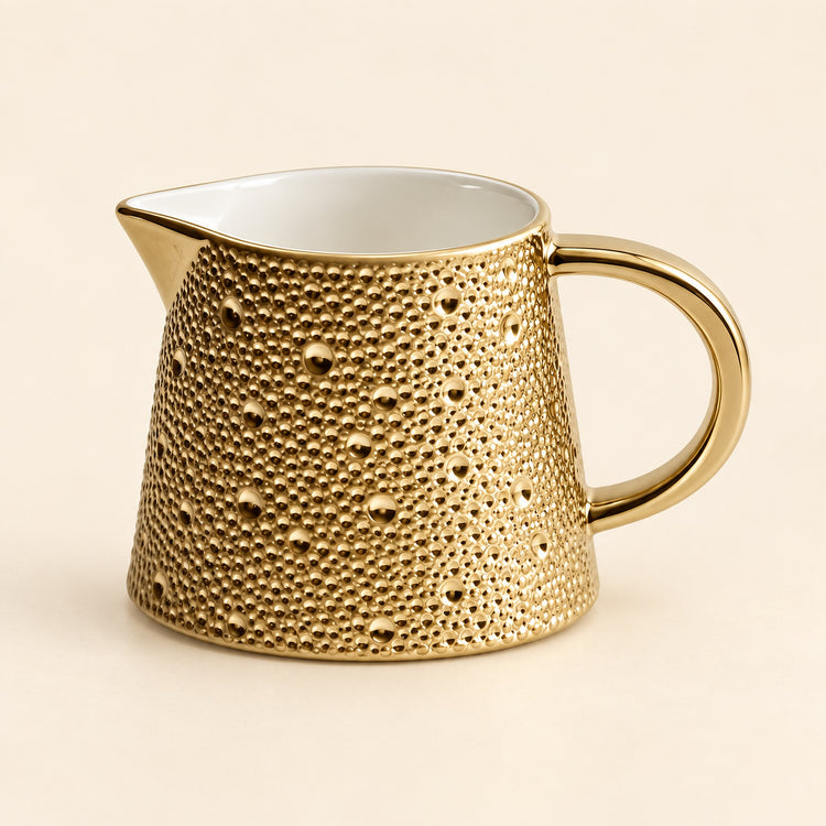 A small gold ceramic pitcher with a textured, dimpled exterior and a smooth white interior, set against a solid light beige background.