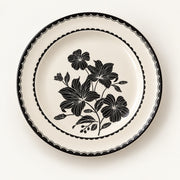 A round ceramic plate with a cream-colored glaze, featuring a black floral pattern in the center and a decorative black scalloped border around the rim, shown against a plain off-white background.