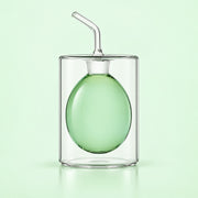 A clear glass double-walled cup with a matching clear glass straw, featuring a translucent light green inner balloon or sphere, set against a solid pale green background.