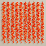 Ten vertical strands of artificial maple leaves in shades of orange, red, and yellow hang against a plain light beige wall.