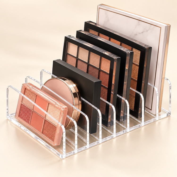 Clear acrylic makeup organizer with multiple compartments holding eyeshadow palettes and a round compact, photographed on a warm beige studio background.