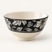 A ceramic bowl with a cream-colored interior and base, featuring a black exterior decorated with a white floral pattern and a scalloped border.
