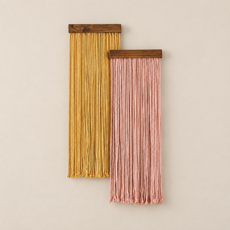 Two vertical macrame wall hangings with wooden tops, one in mustard yellow and one in dusty pink, displayed against a warm beige background.