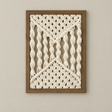 A framed macrame wall art piece with cream-colored woven cords and a medium brown wooden frame, displayed against a neutral beige wall.