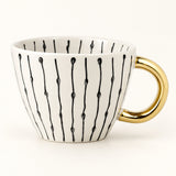 A white ceramic coffee mug with a gold handle and black vertical line patterns, photographed on a clean white background.
