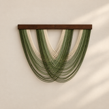 A modern wall hanging sculpture featuring multiple strands of green and cream cord draped from a dark brown wooden bar, mounted on a warm beige wall with soft shadow overlays.