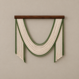 A bohemian-style macrame wall hanging with a dark brown wooden dowel, featuring cream-colored cords and green stripes arranged in a U-shape with fringe details, mounted on a neutral beige wall.