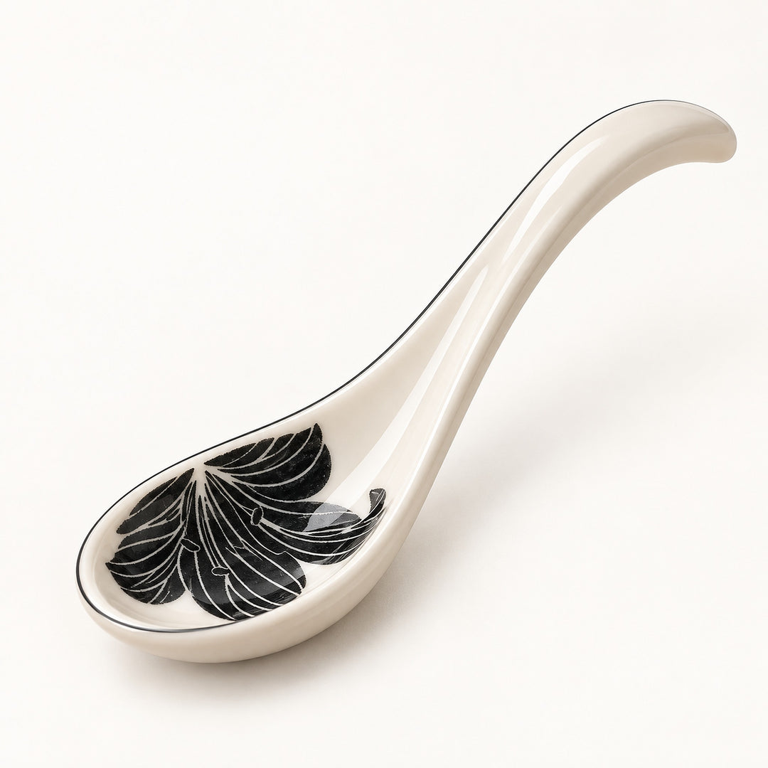 A glossy ceramic serving spoon in an off-white color, featuring a black and white leaf pattern inside the bowl and a thin black rim, set against a plain white background.