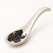 A glossy ceramic serving spoon in an off-white color, featuring a black and white leaf pattern inside the bowl and a thin black rim, set against a plain white background.