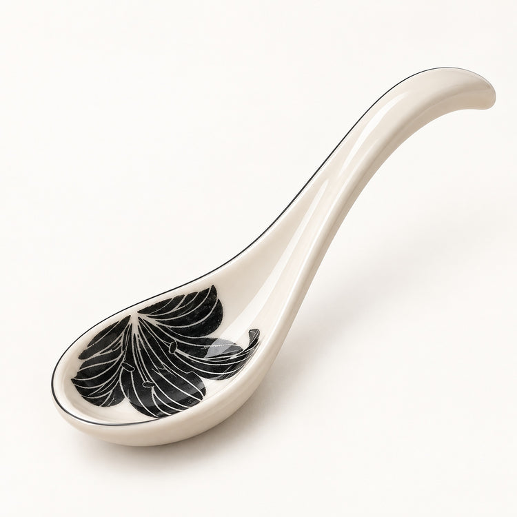 A glossy ceramic serving spoon in an off-white color, featuring a black and white leaf pattern inside the bowl and a thin black rim, set against a plain white background.