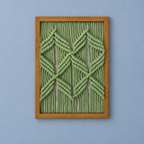 A framed geometric wall art piece featuring knotted sage green rope in a diamond and chevron pattern, enclosed in a light brown wooden frame against a solid periwinkle blue background.