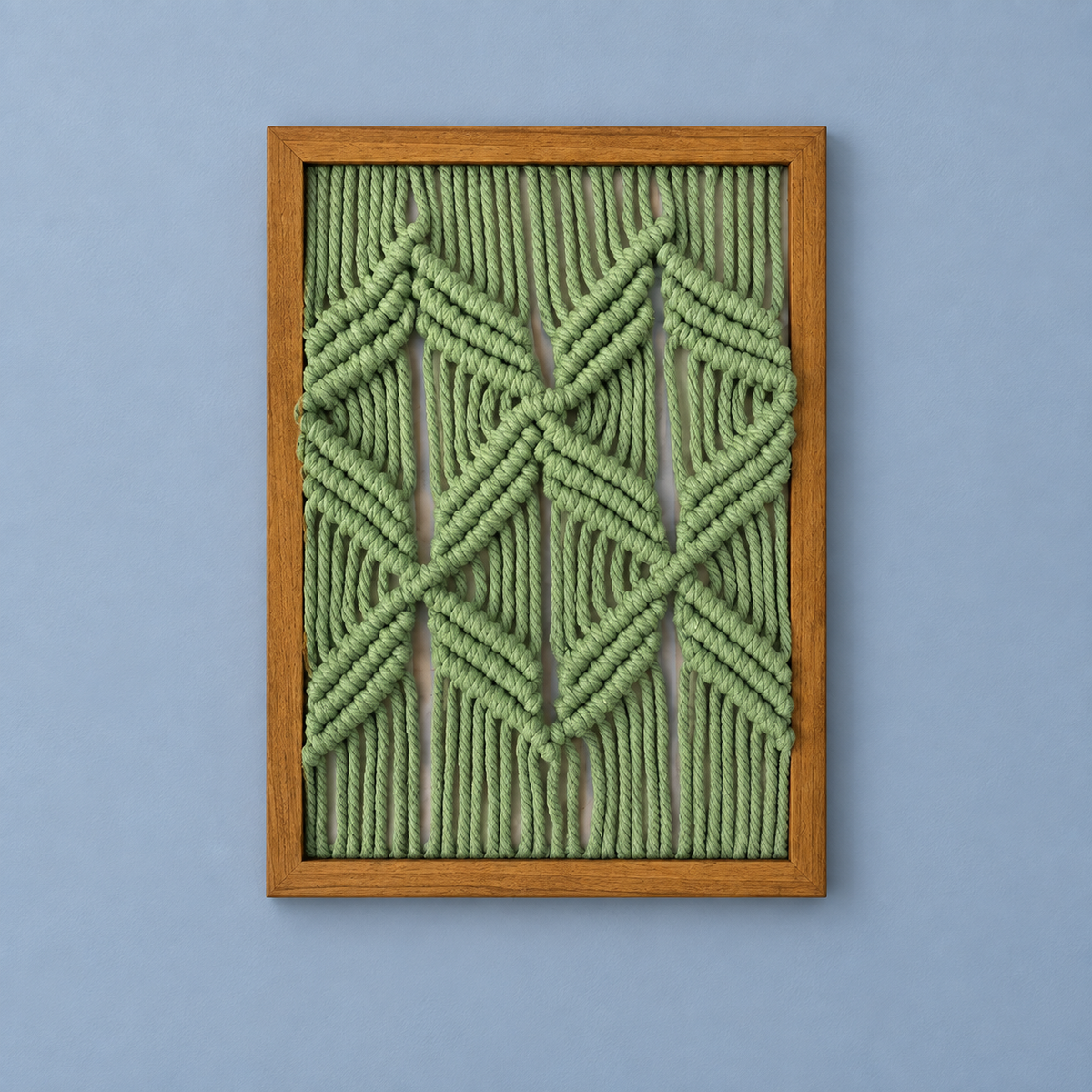 A framed geometric wall art piece featuring knotted sage green rope in a diamond and chevron pattern, enclosed in a light brown wooden frame against a solid periwinkle blue background.
