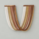 A U-shaped wall art piece made of many hanging strands in white, beige, tan, brown, and mustard yellow, suspended from a wooden top bar against a light gray wall.
