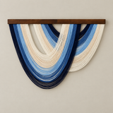 A modern wall hanging sculpture featuring layered, semi-circular strands of dark navy, medium blue, and cream-colored threads suspended from a dark brown wooden bar, mounted on a neutral beige wall.
