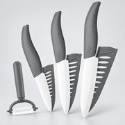 Four kitchen knives and a peeler with dark gray handles and white blades, arranged upright on a light gray studio background.