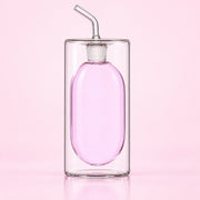 A clear glass double-walled bottle with an oval-shaped pink interior and a curved glass straw, set against a solid light pink background.