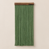A green fringed wall hanging with a dark wood rod at the top, shown against a plain beige wall.