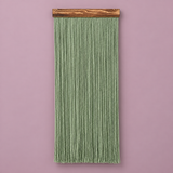 A vertical sage green macrame wall hanging with a natural wood dowel, displayed against a solid dusty pink background.
