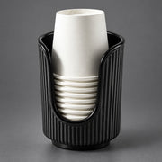 A black ribbed cup holder or cup dispenser holding a stack of white paper cups, photographed on a smooth gray studio background.