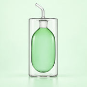 A clear glass double-walled tumbler with a green glass bottle insert and a clear glass angled straw, set against a solid pale green background.