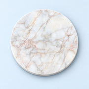 A round white marble board with natural beige and light brown veining, photographed from directly above on a light blue background.