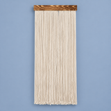 A vertical macrame wall hanging made of thick cream-colored cotton cords suspended from a rectangular dark brown wooden bar, shown against a solid light blue background.