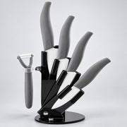 A 5-piece kitchen knife set with gray handles and black blades, displayed on a black magnetic stand with a matching gray peeler to the left, set against a light gray studio background.