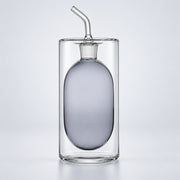 A clear glass double-walled bottle with a frosted oval center and a curved glass straw on a light gray background.