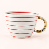 A white ceramic coffee mug with thin red horizontal stripes and a polished gold handle, set against a solid pale pink background.