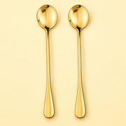 Two long gold spoons with oval bowls and tapered handles, shown upright on a warm cream background.