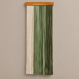 A vertical macrame wall hanging with a natural wood top bar, featuring three vertical panels of woven cord in cream, sage green, and olive green, shown against a solid warm gray background.