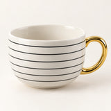 A white ceramic coffee mug with thin black horizontal stripes and a shiny gold handle, shown on a clean off-white background.