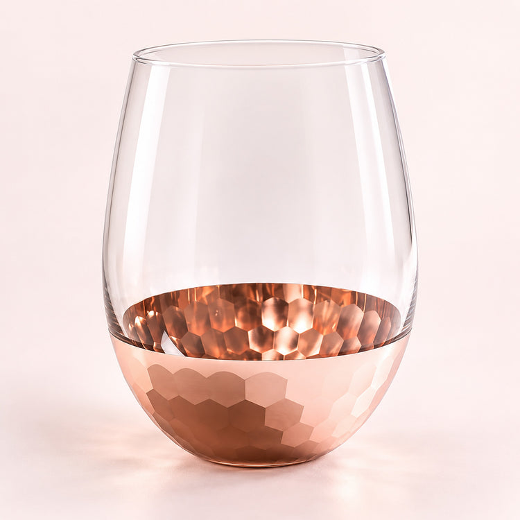 A stemless wine glass with a hammered copper-colored metallic base, shown on a soft light pink background.