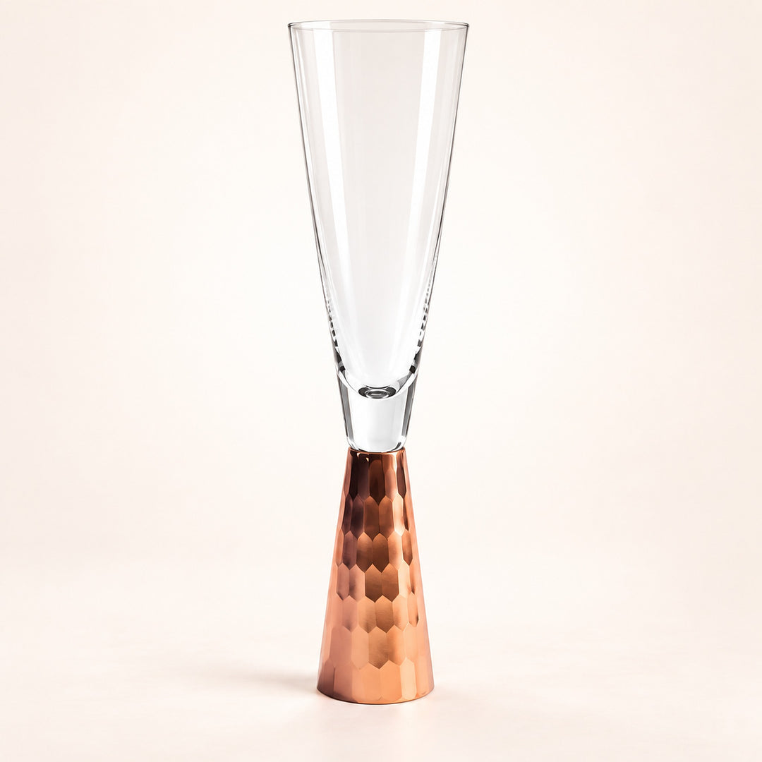 A clear glass champagne flute with a hammered copper-colored metal base, photographed against a clean off-white studio background.