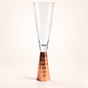 A clear glass champagne flute with a hammered copper-colored metal base, photographed against a clean off-white studio background.