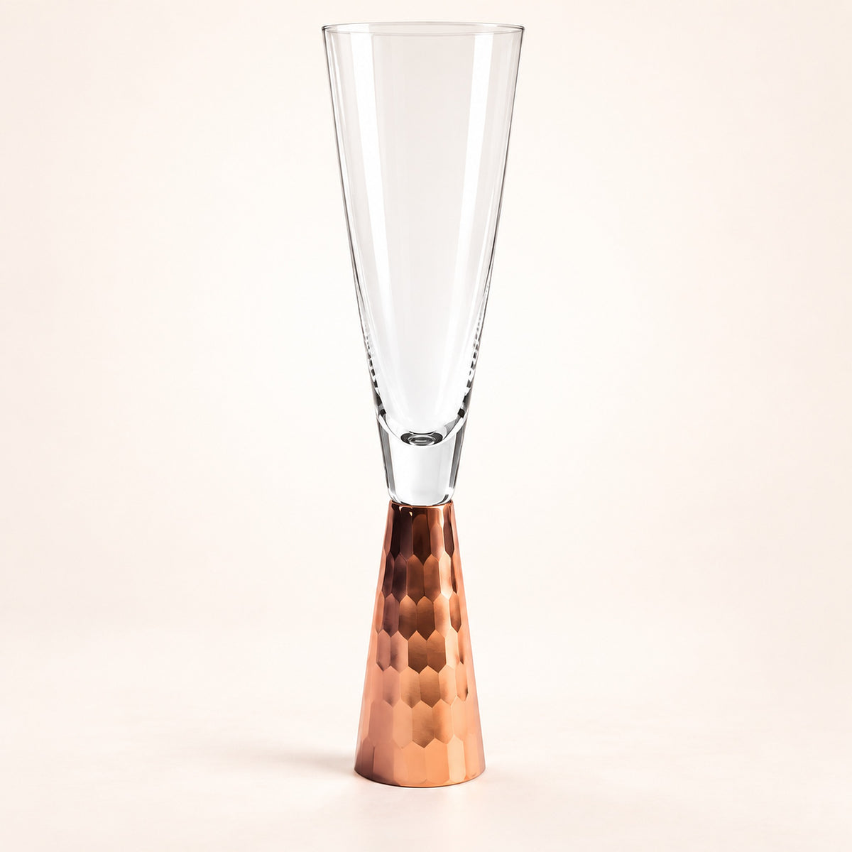 A clear glass champagne flute with a hammered copper-colored metal base, photographed against a clean off-white studio background.