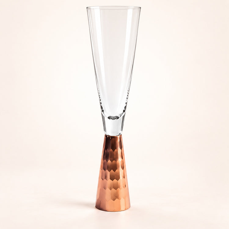 A clear glass champagne flute with a hammered copper-colored metal base, photographed against a clean off-white studio background.