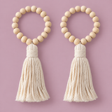 Two matching beaded tassel keychains on a solid dusty pink background. Each keychain features a circle of light natural wood beads with a thick cream-colored cotton rope tassel hanging below.