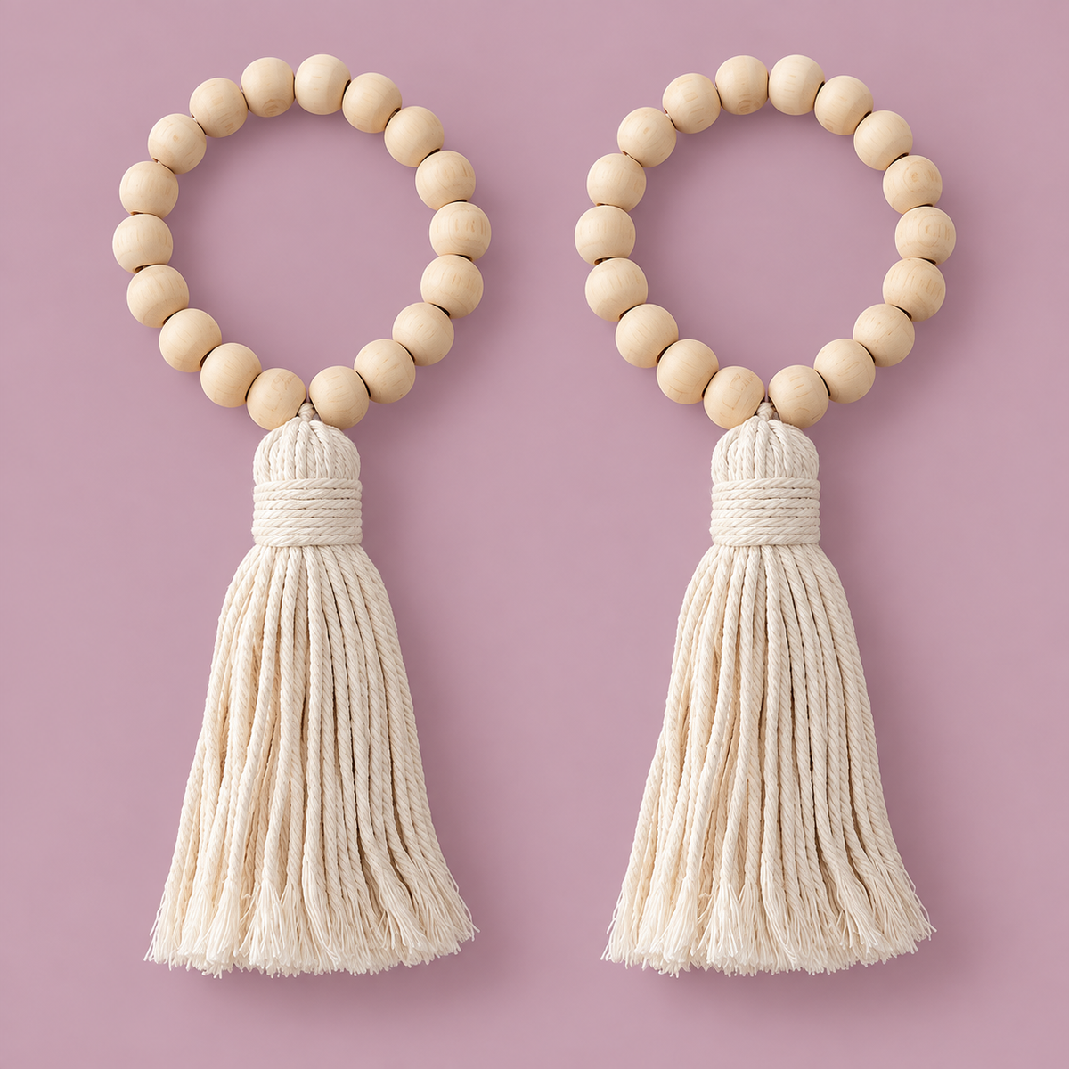 Two matching beaded tassel keychains on a solid dusty pink background. Each keychain features a circle of light natural wood beads with a thick cream-colored cotton rope tassel hanging below.