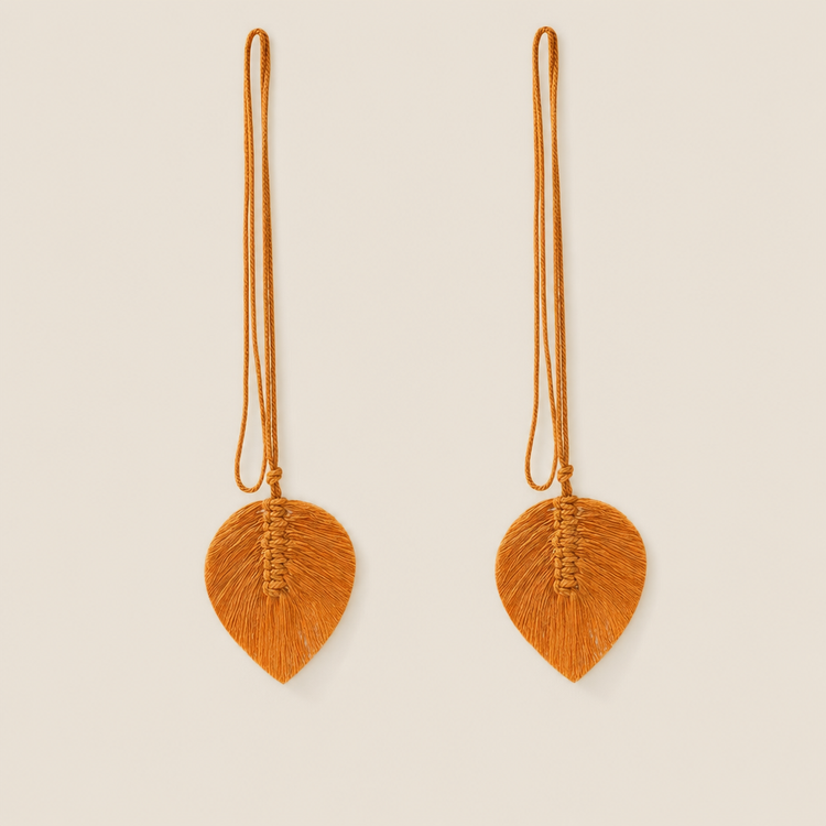 A pair of long, leaf-shaped orange tassel earrings suspended from thin cords, displayed against a solid light beige background.