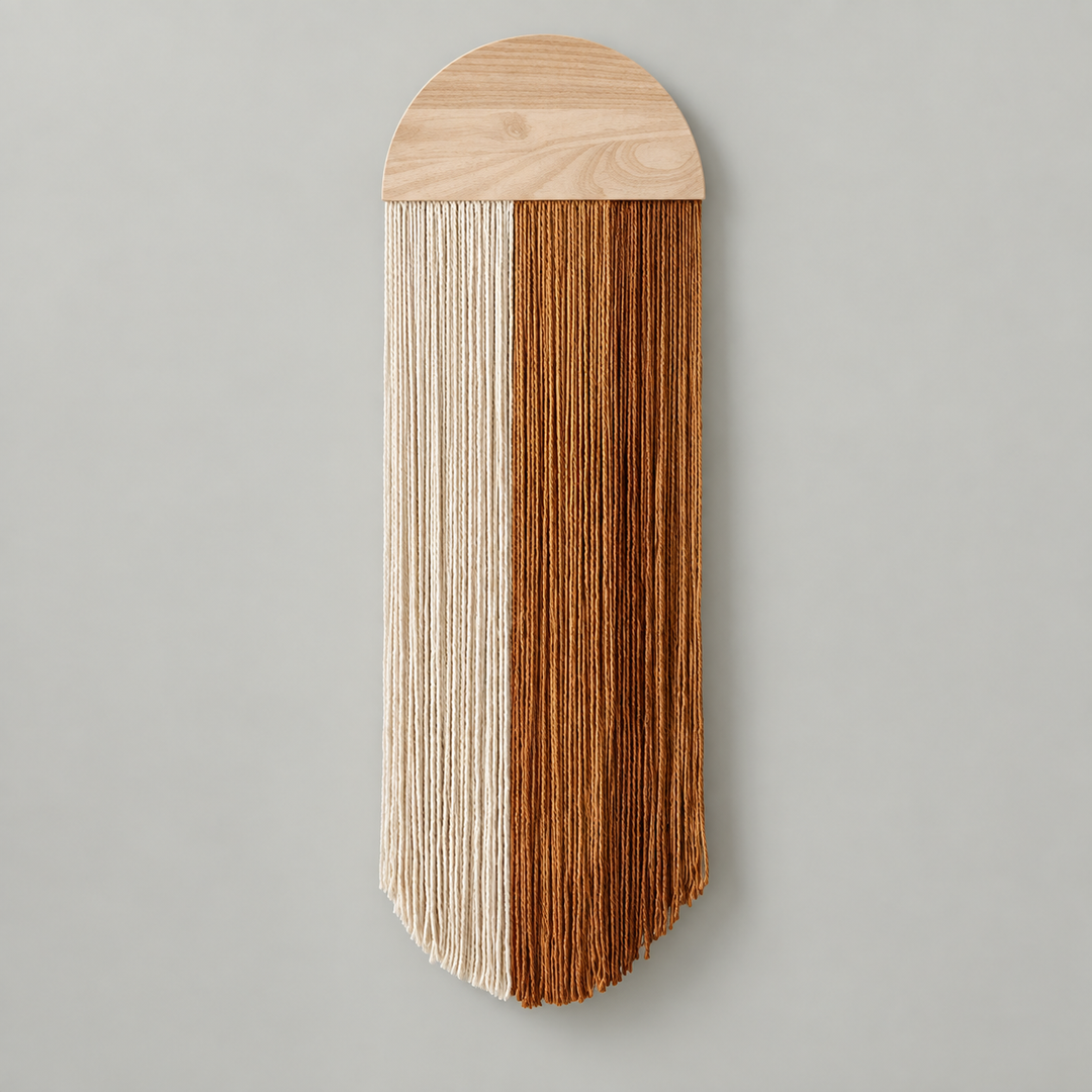A vertical wall hanging with a light natural wood top panel and long fringe strands split into two vertical sections: one in off-white and one in warm medium brown, shown against a plain light gray wall.