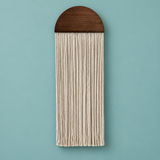 A vertical wall hanging with a dark brown wooden semicircular top and long cream-colored fringe strands, shown against a solid light blue background.
