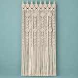 A cream macrame wall hanging with intricate woven patterns and long fringe, displayed against a solid teal background.