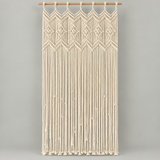 Cream macrame wall hanging with geometric woven patterns and long fringe, suspended from a wooden rod against a light gray background.
