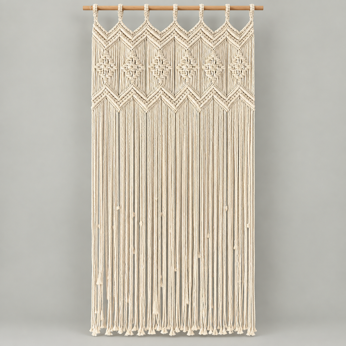 Cream macrame wall hanging with geometric woven patterns and long fringe, suspended from a wooden rod against a light gray background.