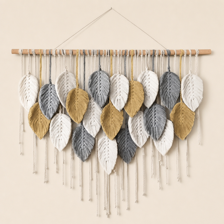 A handmade macrame wall hanging featuring knitted leaf shapes in white, light gray, and mustard yellow, suspended from a light wood dowel against a warm beige wall.