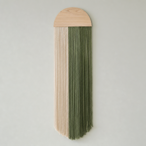 A vertical wall hanging with a light natural wood semicircular top and long fringe strands in two vertical sections: one in pale cream and one in muted sage green, shown against a plain light gray wall.